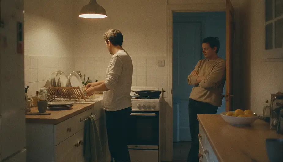 Two adults in a small kitchen under one pendant lamp in the evening, one at the draining board seen from behind and one standing in the doorway with arms folded.