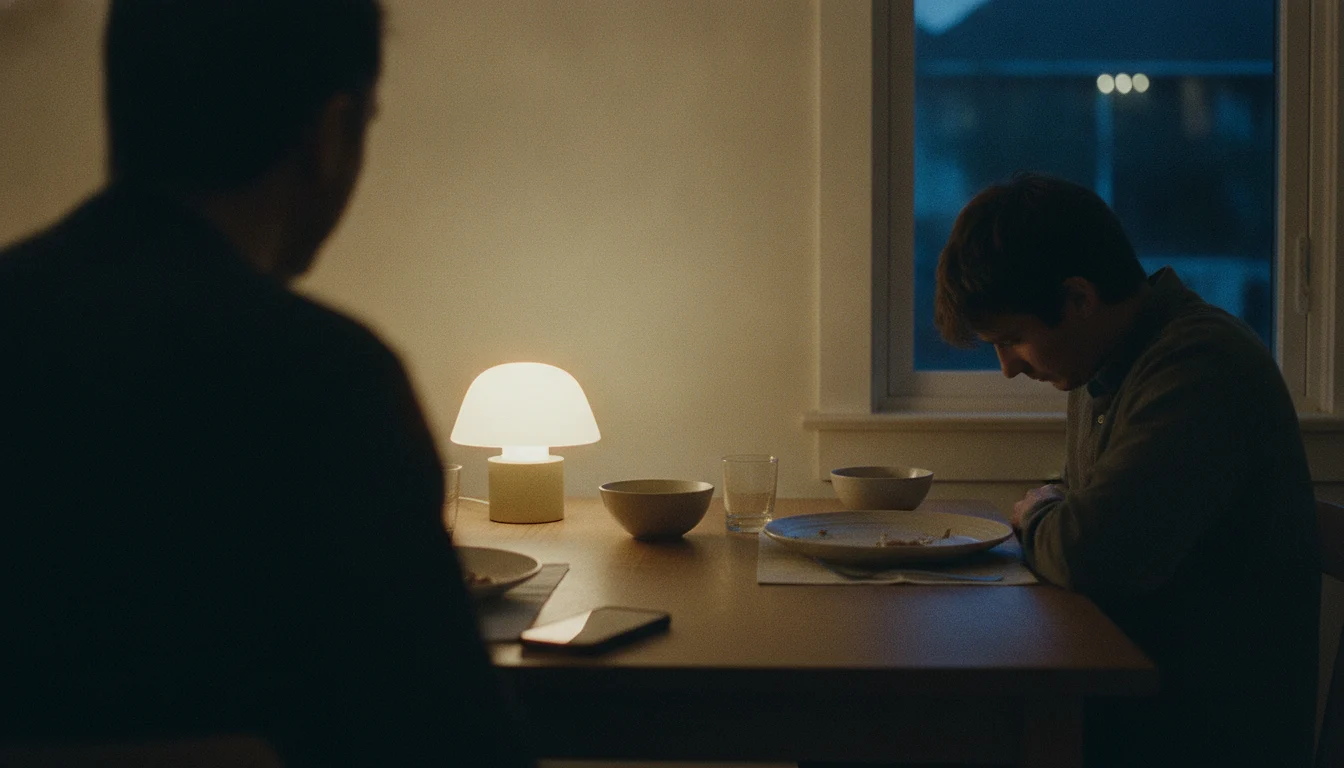A dinner table in the evening seen past the dark shoulder of one person; a phone lies on the wood, the other person sits with their head lowered.